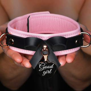 Pink and black bondage bow collar with rose gold love heart 'Good girl' tag held in hands against a blurred background